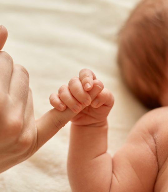Faceless mother with naked baby, infant holding mommy's finger, mum spending time with her tiny child on light background.
